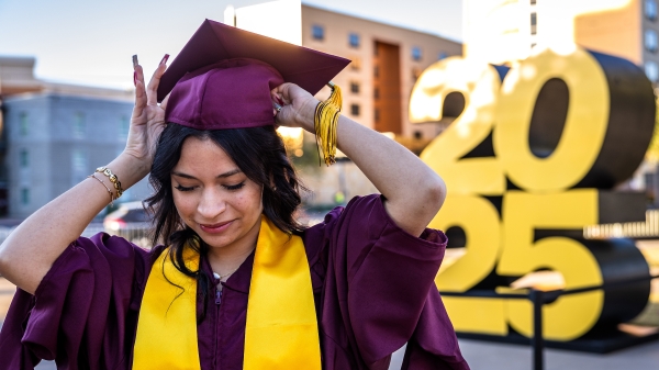 A woman in a maroon graduation cap and gown adjusts her cap in front of a large 2025 display sign