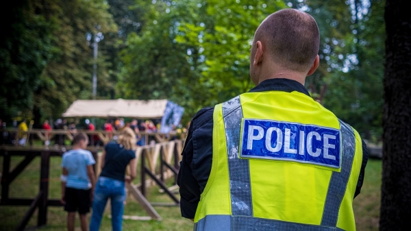 Man wearing a bright vest that reads "police" seen from behind looking toward people at an event.