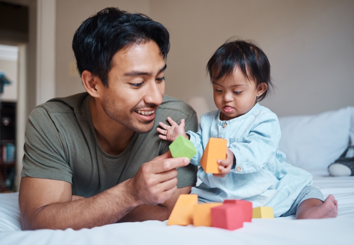 Baby with Down syndrome and father playing with educational blocks in a bedroom.